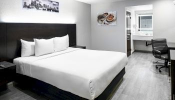 A modern hotel room features a large bed, artwork on the walls, a desk with a chair, and a bathroom with a sink and mirror visible in the background.