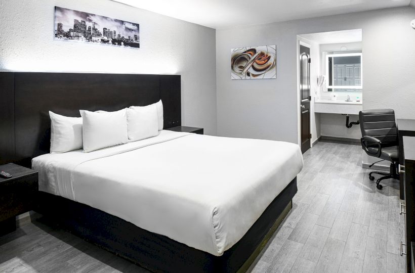A modern hotel room features a large bed, artwork on the walls, a desk with a chair, and a bathroom with a sink and mirror visible in the background.