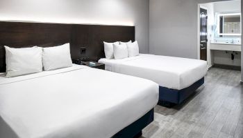 A hotel room features two double beds with white linens, a modern decor, bedside tables, and an adjacent bathroom with a sink and mirror.