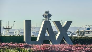 The image shows large "LAX" letters with an airport control tower in the background and pink flowers in the foreground.