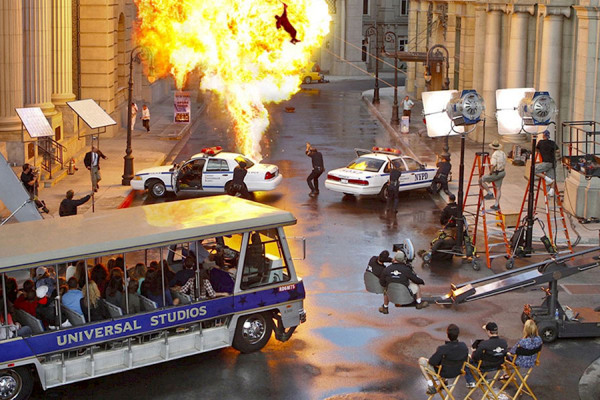 A film set with an explosion, police cars, crew, and a Universal Studios tour tram filled with spectators can be seen.