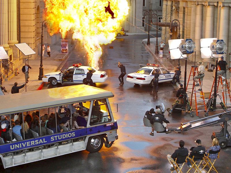 A film set with an explosion, police cars, crew, and a Universal Studios tour tram filled with spectators can be seen.