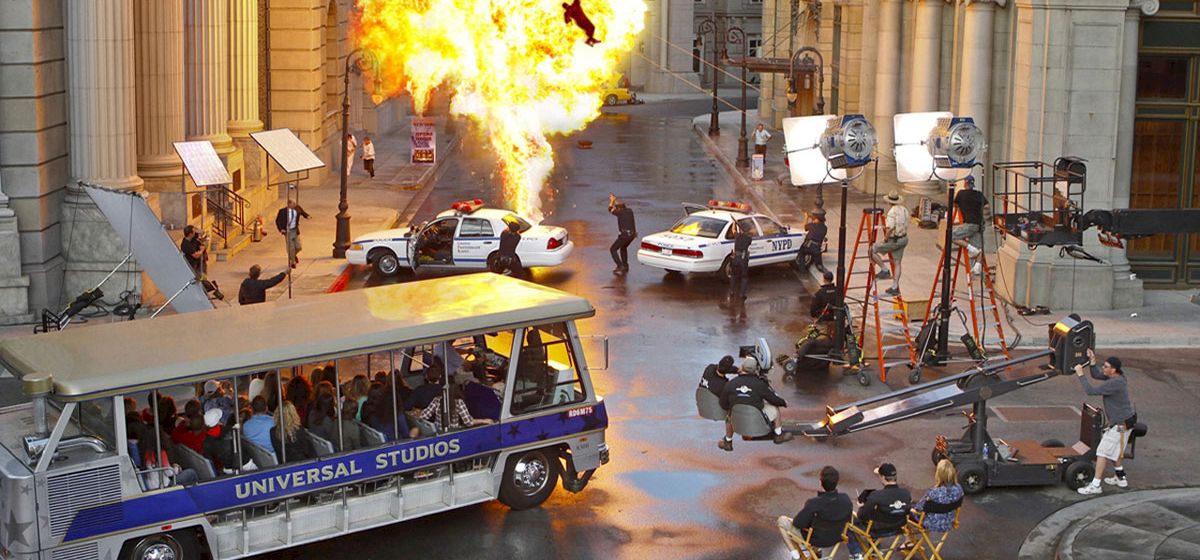 A film set with an explosion, police cars, crew, and a Universal Studios tour tram filled with spectators can be seen.