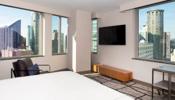 A modern hotel room with city views, a bed, wall-mounted TV, armchair, and small table by the large windows overlooking skyscrapers ending the sentence.