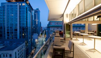 The image shows a modern balcony with tables and chairs, overlooking a cityscape with tall buildings and a street below during dusk or early evening.