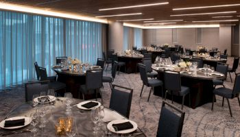The image shows an elegant dining setup in a banquet hall, with round tables set with black napkins, glassware, and floral centerpieces, under modern lighting.