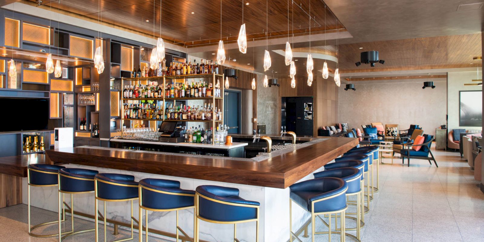 A modern bar with a wooden counter, blue high stools, a well-stocked back wall, and stylish pendant lights; seating area in the background.