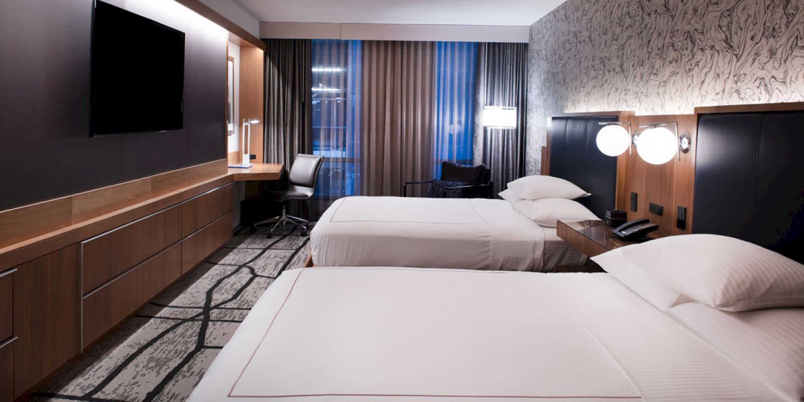 A modern hotel room with two neatly made beds, a wall-mounted TV, a desk by the window, and a stylish decor with patterned walls and carpet.
