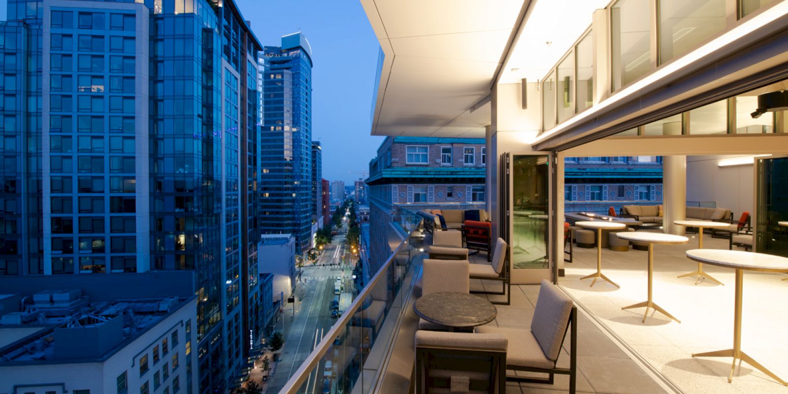A modern rooftop terrace with seating areas and tables overlooks a city street lined with tall buildings at dusk, featuring a vibrant urban scene.