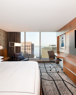A modern hotel room features a large bed, a desk with a chair, wall art, a large TV, and a window with a city and water view in the distance.