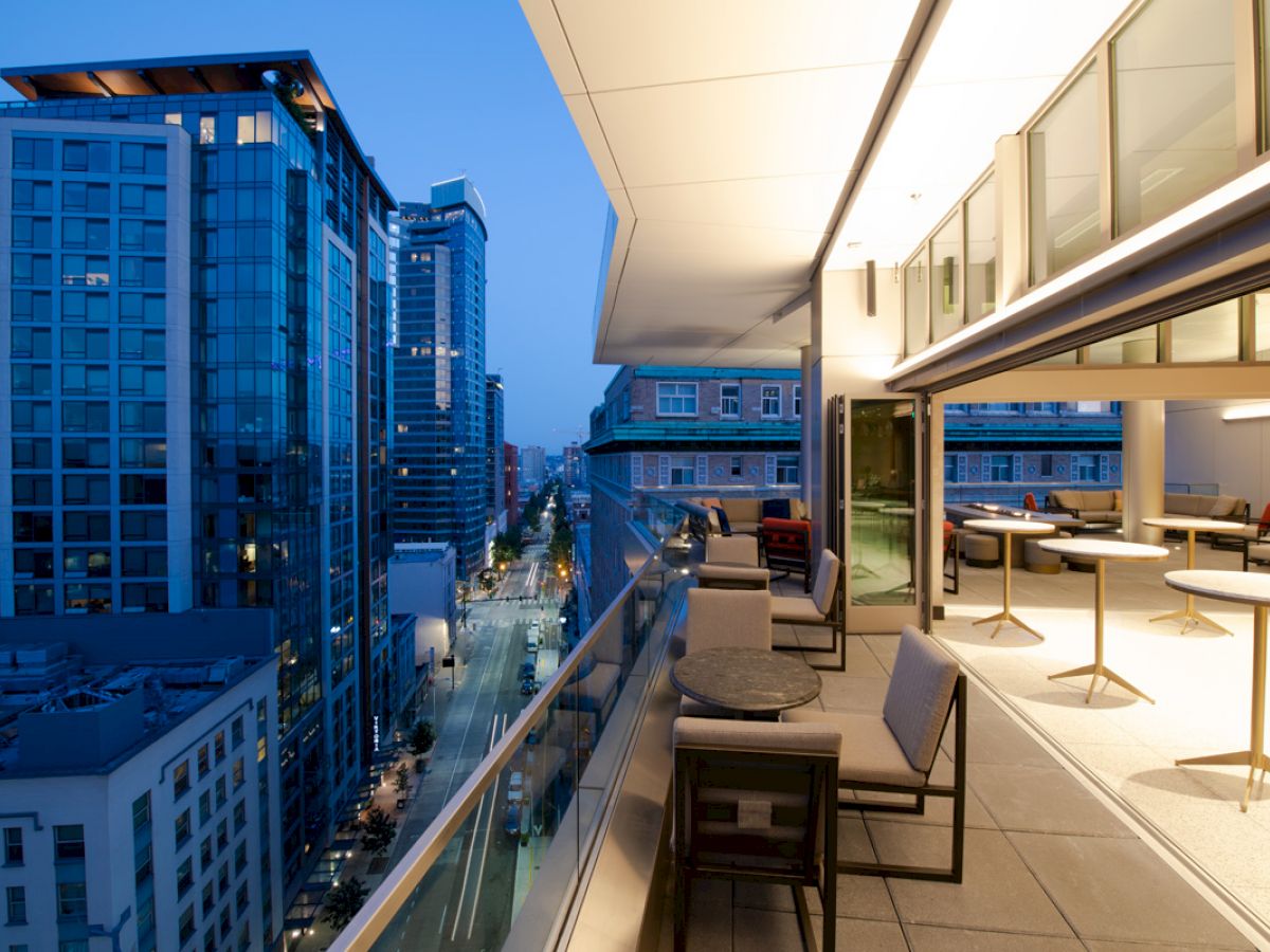 A modern balcony view at dusk, featuring city skyscrapers and a spacious outdoor seating area with tables and chairs.