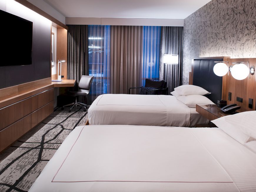 A modern hotel room with two beds, a wall-mounted TV, desk, chair, and a window with curtains letting in natural light, creating a cozy atmosphere.