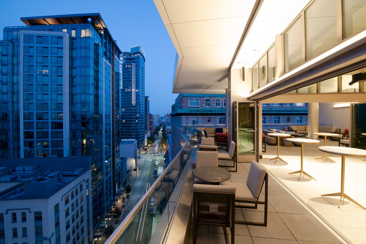 A modern rooftop terrace at twilight, featuring tables and chairs, overlooking a city street lined with tall buildings.