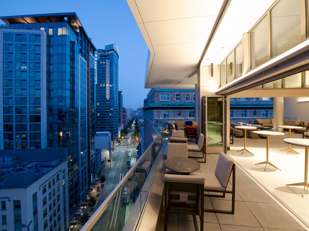 A modern rooftop terrace at twilight, featuring tables and chairs, overlooking a city street lined with tall buildings.