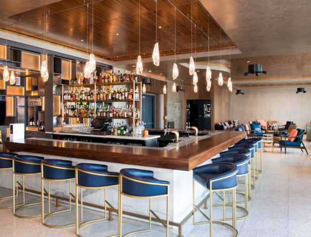 This image shows a modern bar with blue barstools, hanging lights, and a well-stocked shelf of liquor bottles. There are tables in the background.