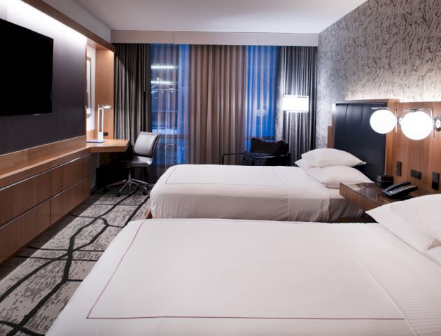 A modern hotel room with two beds, a desk with a chair, a large TV, and stylish decor. The room is well-lit and inviting.