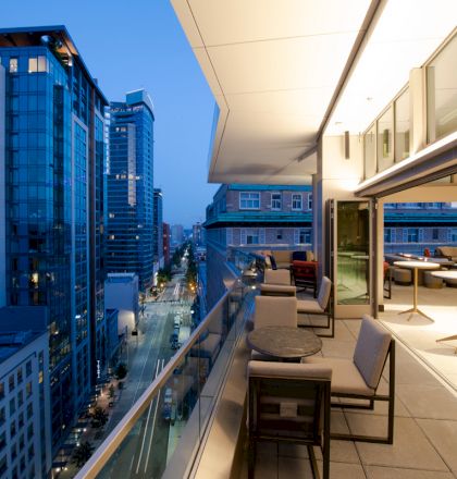 The image shows a modern cityscape at dusk, with tall buildings and a spacious, well-lit balcony featuring tables, chairs, and an open indoor space.