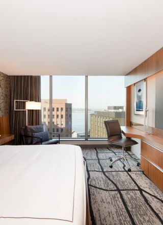 A modern hotel room features a large bed, a wall-mounted TV, a desk with a chair, and a window with a city view.