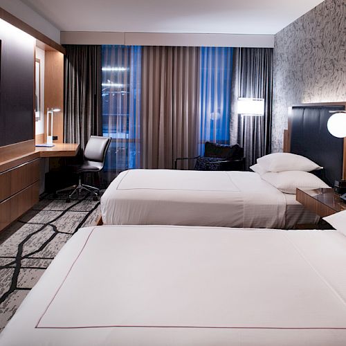 The image shows a modern hotel room with two neatly made beds, a flat-screen TV, a desk, a chair, and a large window with curtains.
