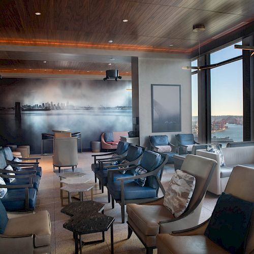 A modern lounge area with plush seating, a mural of a city skyline on the wall, and large windows offering a scenic view of a waterfront and Ferris wheel.