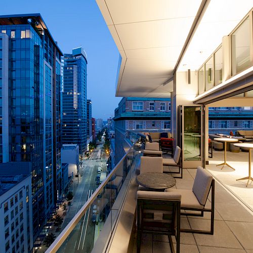 A modern cityscape at dusk with high-rise buildings, a lit balcony with chairs and tables, and a street below.