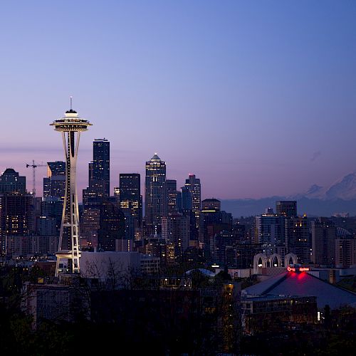A twilight cityscape features numerous buildings and a prominent tower with a mountain in the background, likely in Seattle, Washington.