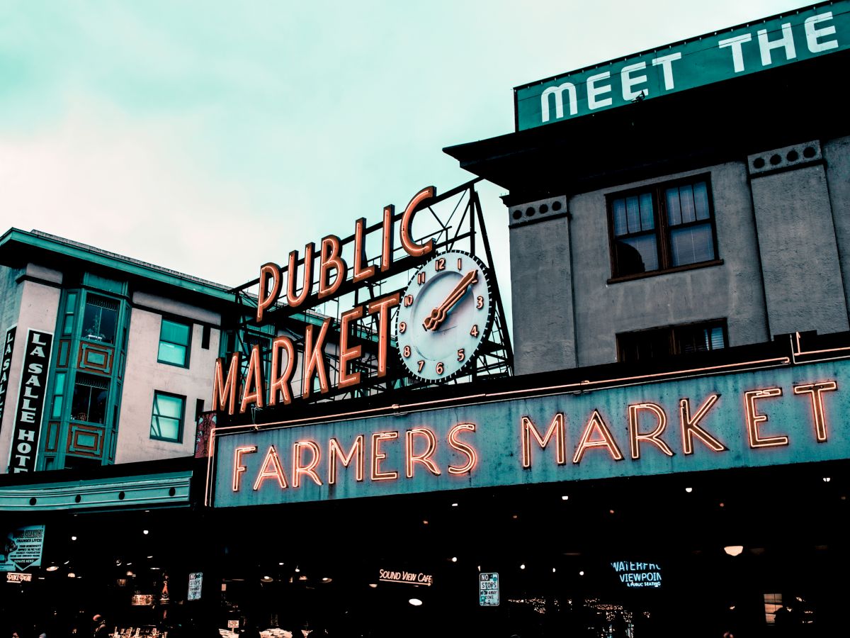 The image shows two buildings with neon signage reading "Public Market" and "Farmers Market" with a clock in between, against a cloudy sky.