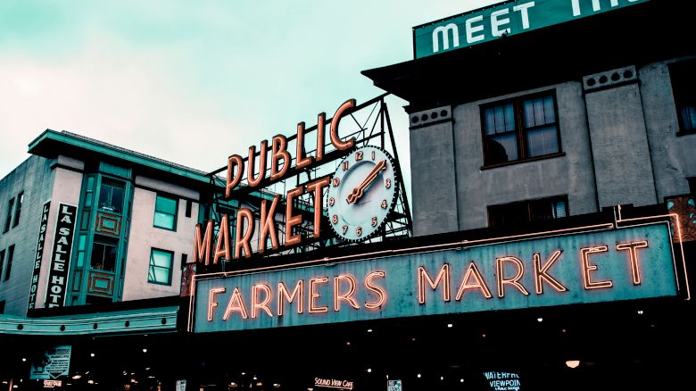 The image shows two buildings with neon signage reading "Public Market" and "Farmers Market" with a clock in between, against a cloudy sky.