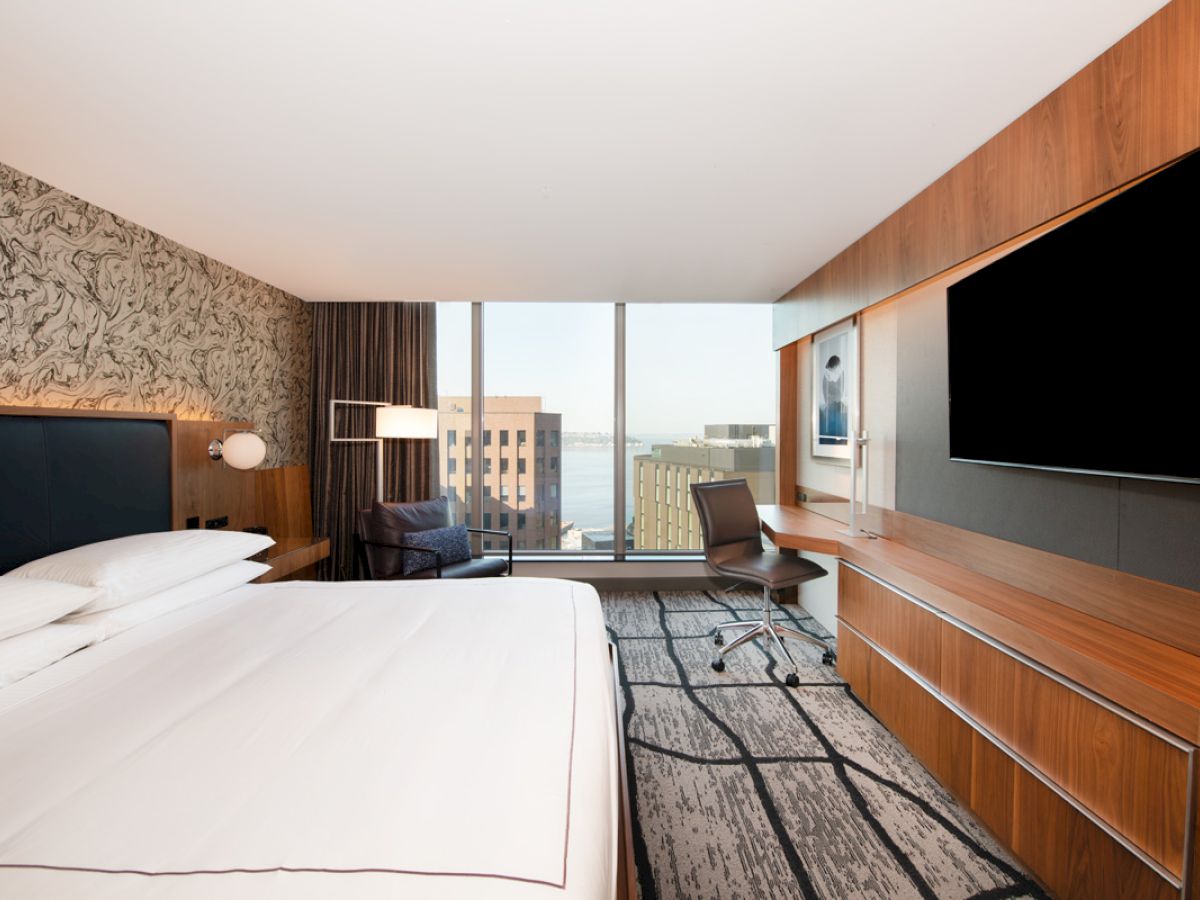 A modern hotel room with a king-sized bed, a large wall-mounted TV, a work desk, a chair by the window, and a view of city buildings outside.