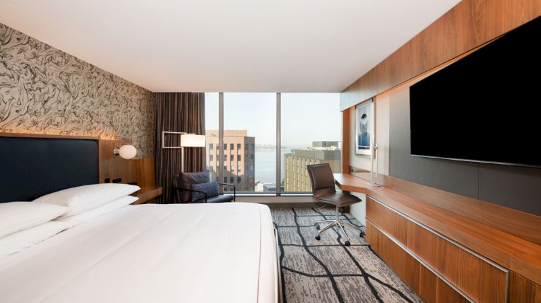 A modern hotel room with a king-sized bed, a large wall-mounted TV, a work desk, a chair by the window, and a view of city buildings outside.