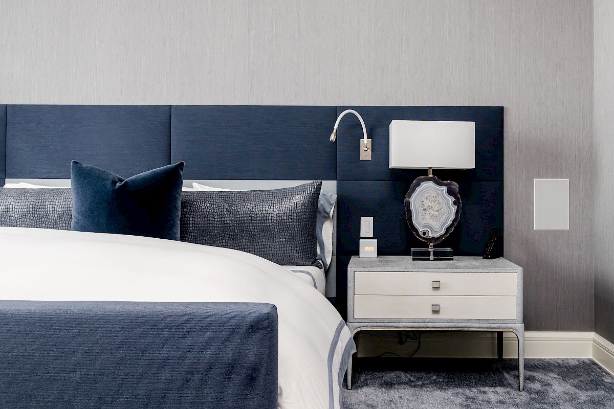 A modern bedroom with a blue upholstered headboard, white and blue bedding, a nightstand with a lamp, and a decorative mirror.