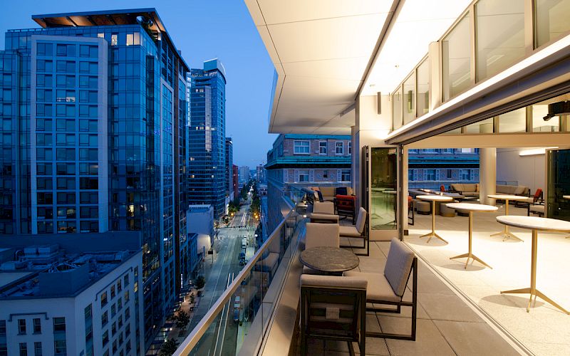 A modern, luxurious balcony with seating overlooks a city street and tall buildings during twilight, showcasing both urban and serene elements.