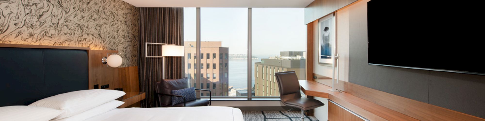 A modern hotel room with a large bed, TV, desk, and floor-to-ceiling windows offering a city and water view.