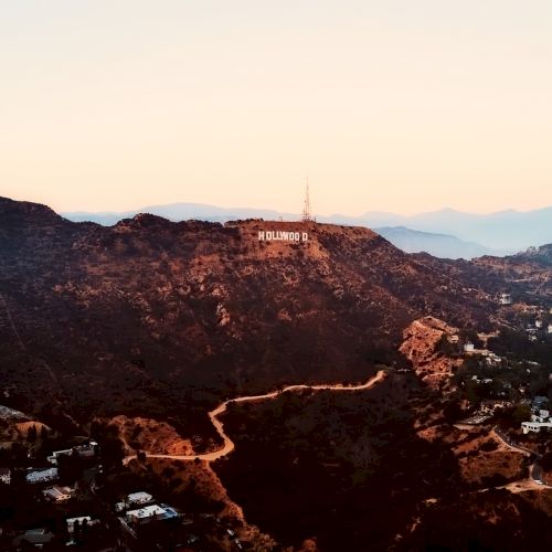 The image shows a mountainous landscape at sunset with a famous white sign on the hill. Various paths and houses are visible scattered across the terrain.