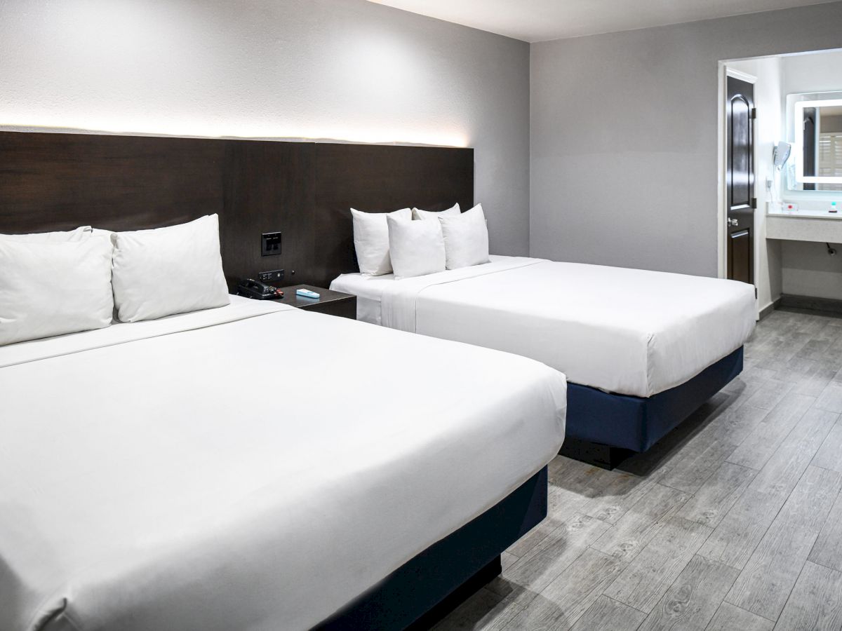 The image shows a hotel room with two neatly made beds, a wooden headboard, and a minimalist design. There's an open bathroom door in the background.