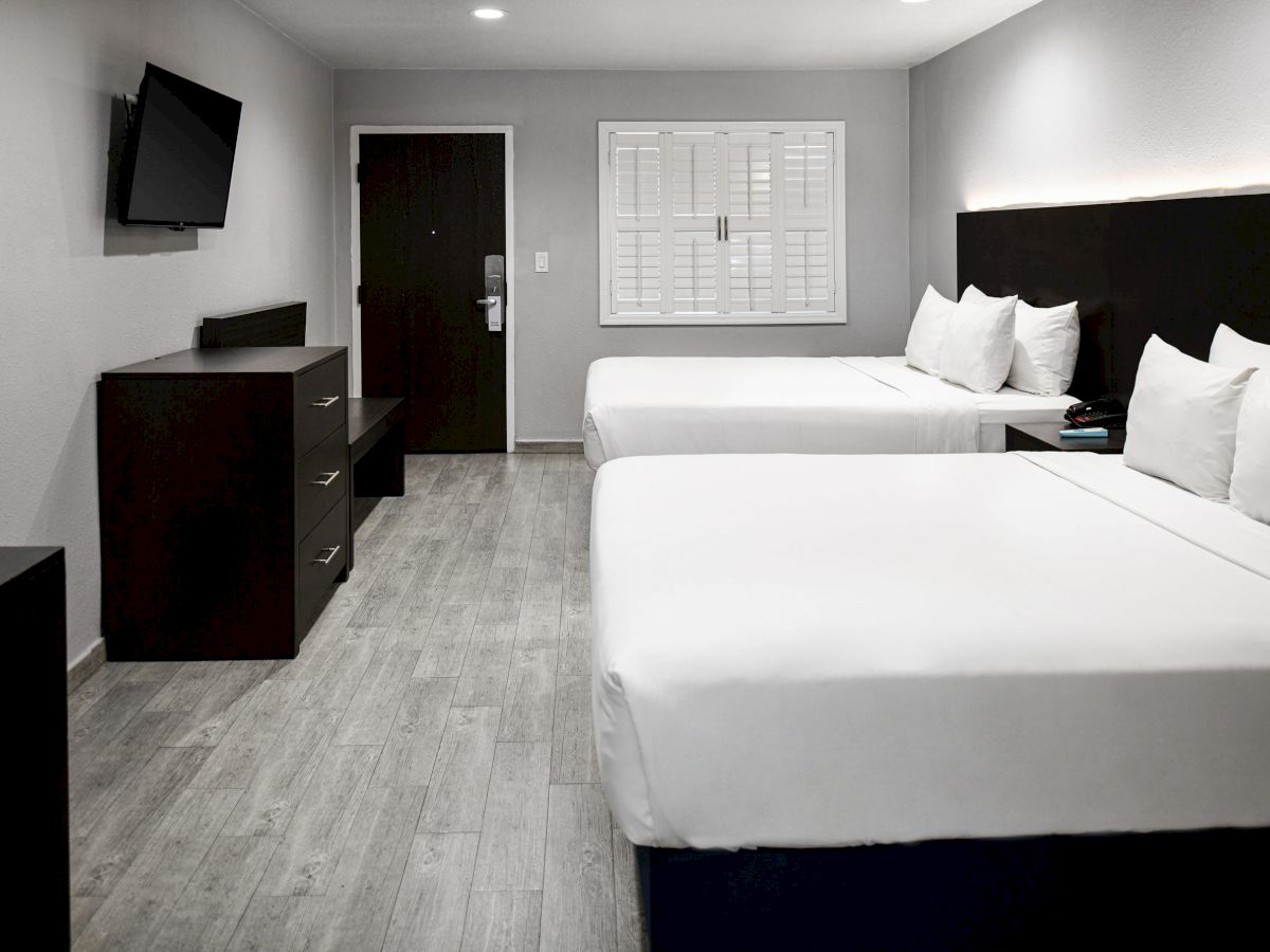 This image shows a modern hotel room with two double beds, a wall-mounted TV, and a dresser under it, all set on a light-colored wood floor.