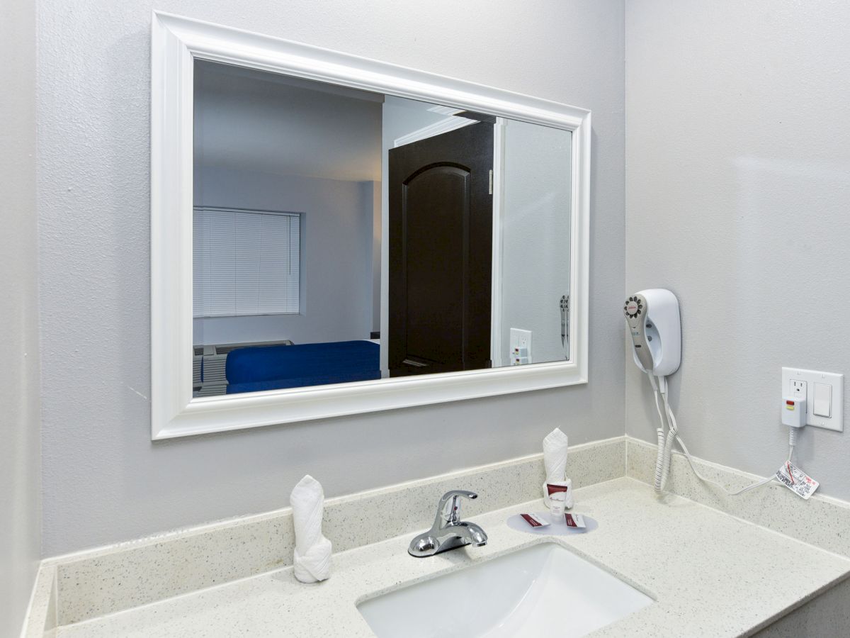 A bathroom sink area with a mirror, a faucet, two wrapped cups, a wall-mounted hairdryer, and a power outlet.