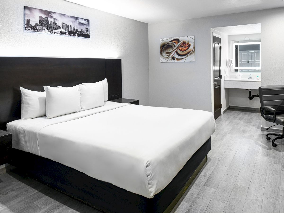 The image shows a modern hotel room with a large bed, wall art, a desk with a chair, and a mirror in the adjacent bathroom area.