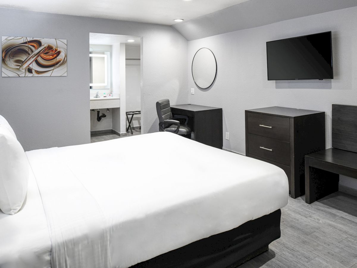 This image shows a modern hotel room with a large bed, desk, chair, TV, dresser, and bathroom. The decor is minimal and clean.